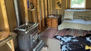 L’intérieur d’un chalet rustique, avec un poêle à bois, un lit recouvert d’une couverture en peau de vache et une fenêtre donnant sur la forêt.