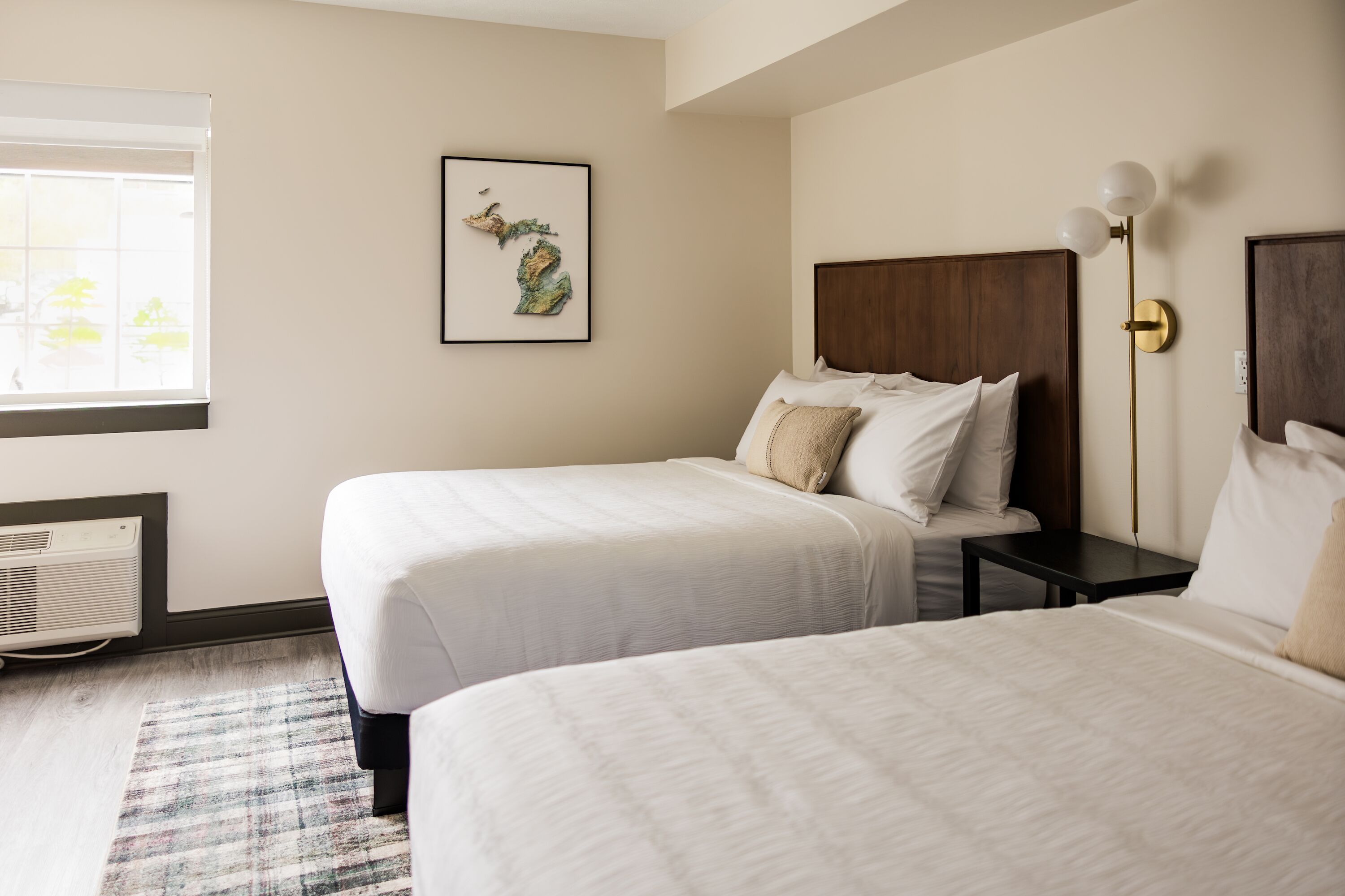 A hotel room with two beds, a framed map on the wall, a bedside table with a lamp, and a window with blinds.