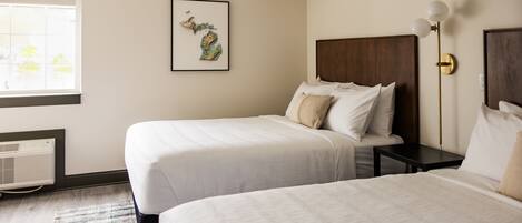 A hotel room with two beds, a framed map on the wall, a bedside table with a lamp, and a window with blinds.