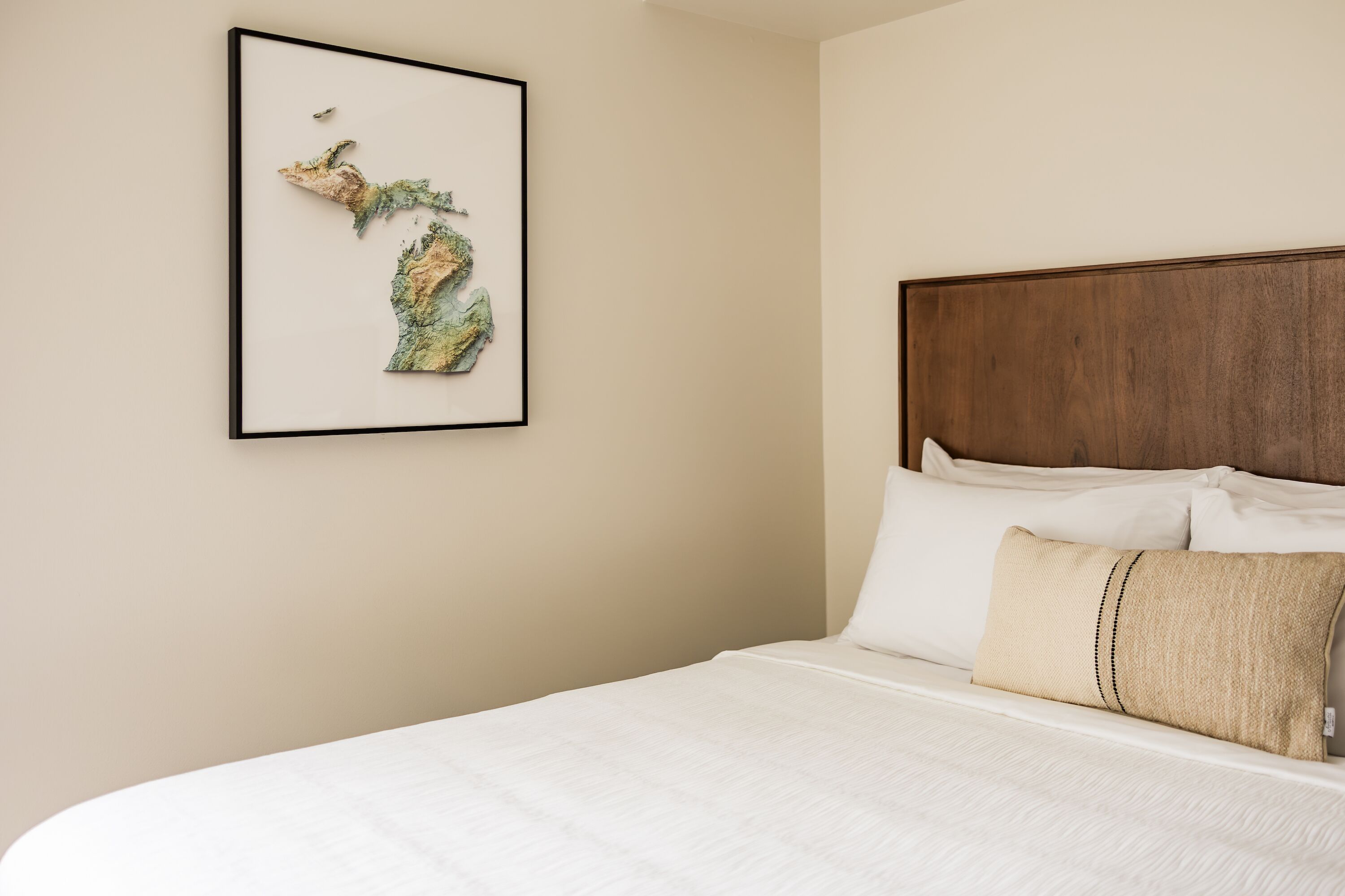 A neatly made bed with white linens and a beige pillow, a framed map on the wall, and a wooden headboard.