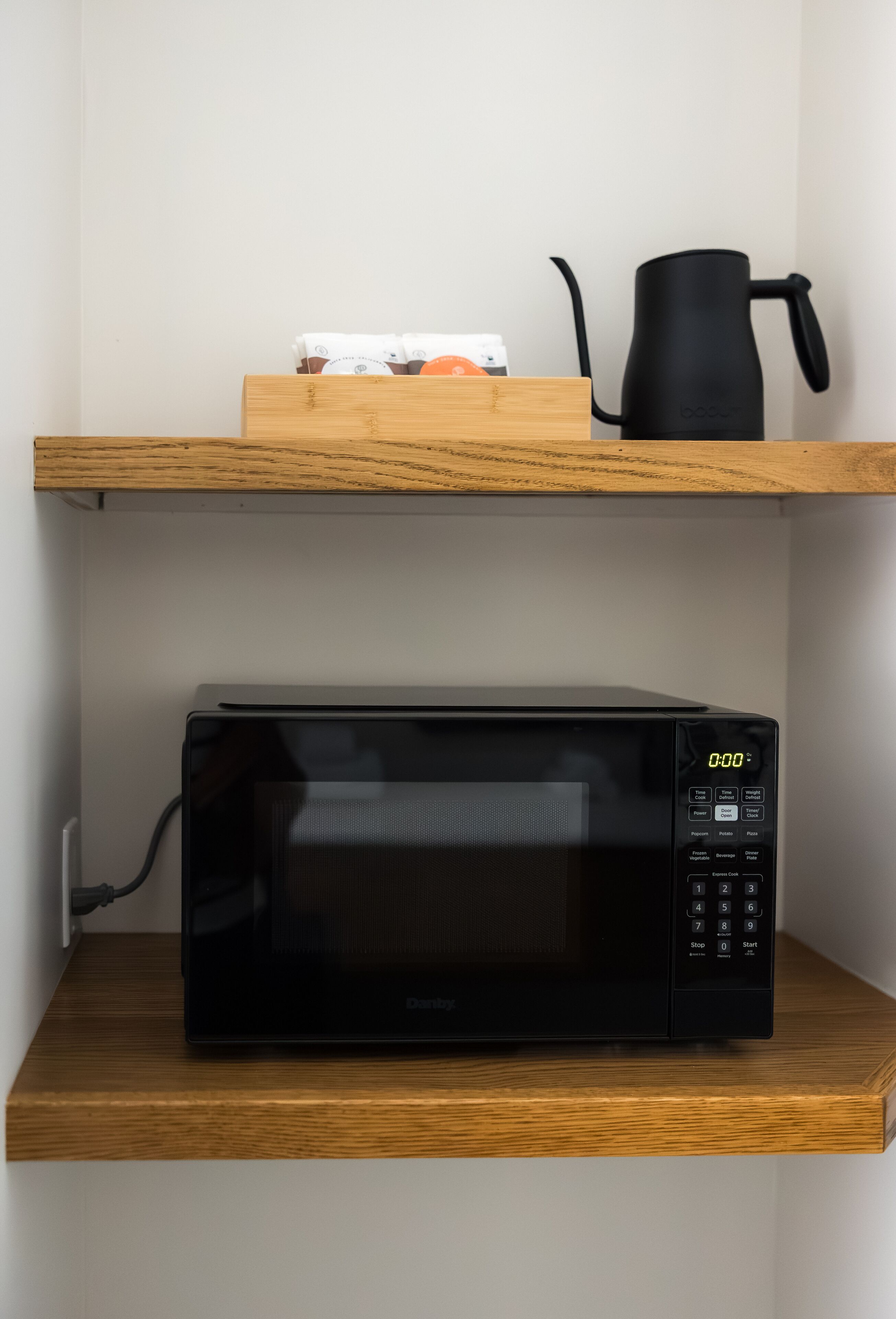 A black microwave on a wooden shelf with a coffee maker and a box of coffee pods above it.