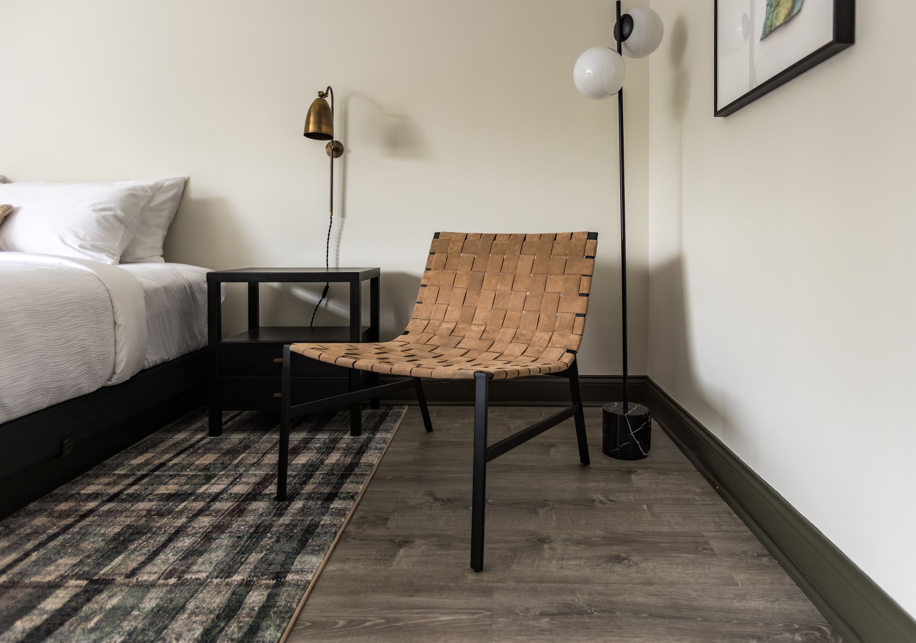A bedroom with a bed, a woven chair, a small table, a lamp, and a picture on the wall.