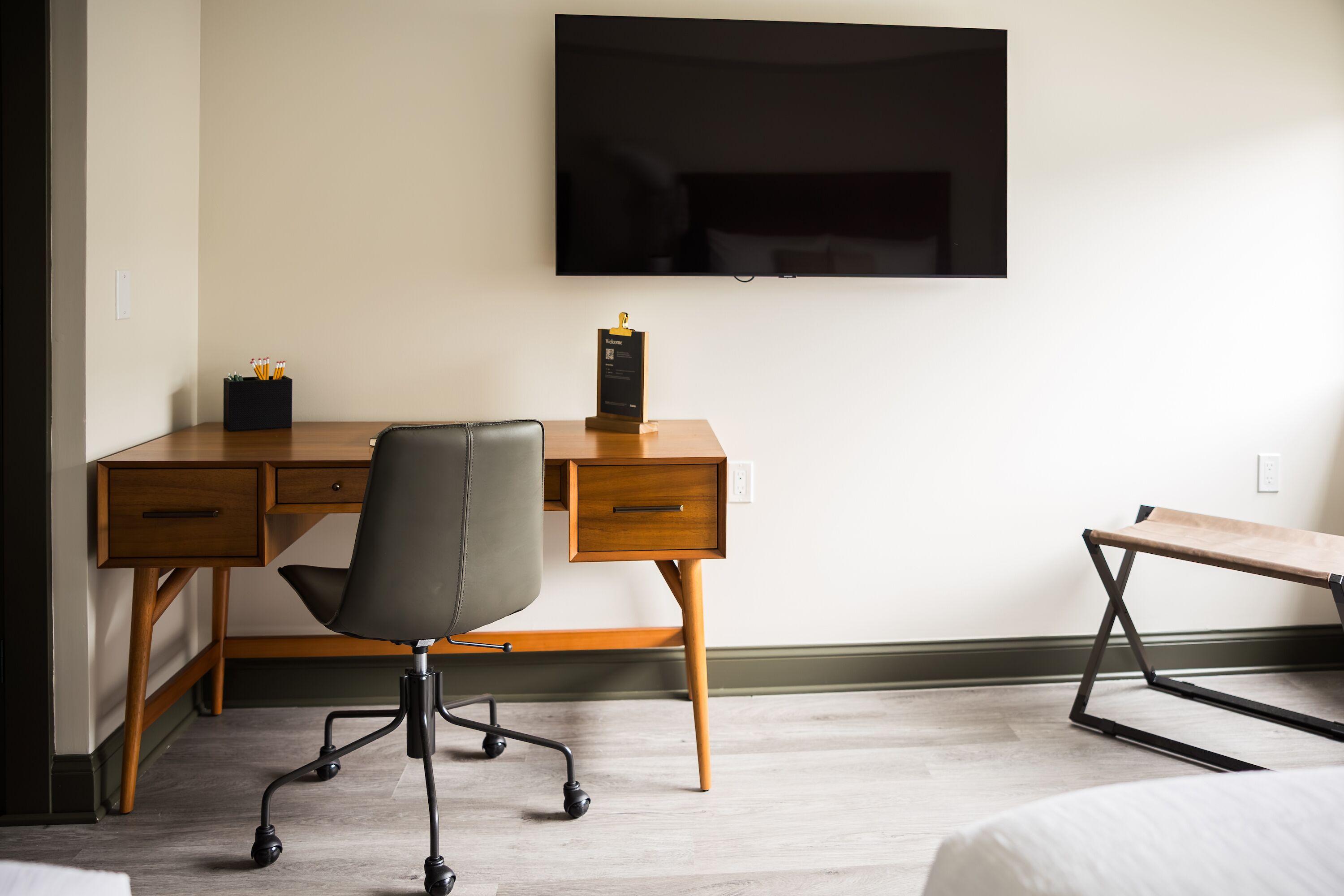 Ein Hotelzimmer mit einem Holztisch, einem schwarzen Bürostuhl, einem an der Wand befestigten Flachbildfernseher und einem Bett mit weißer Bettwäsche.