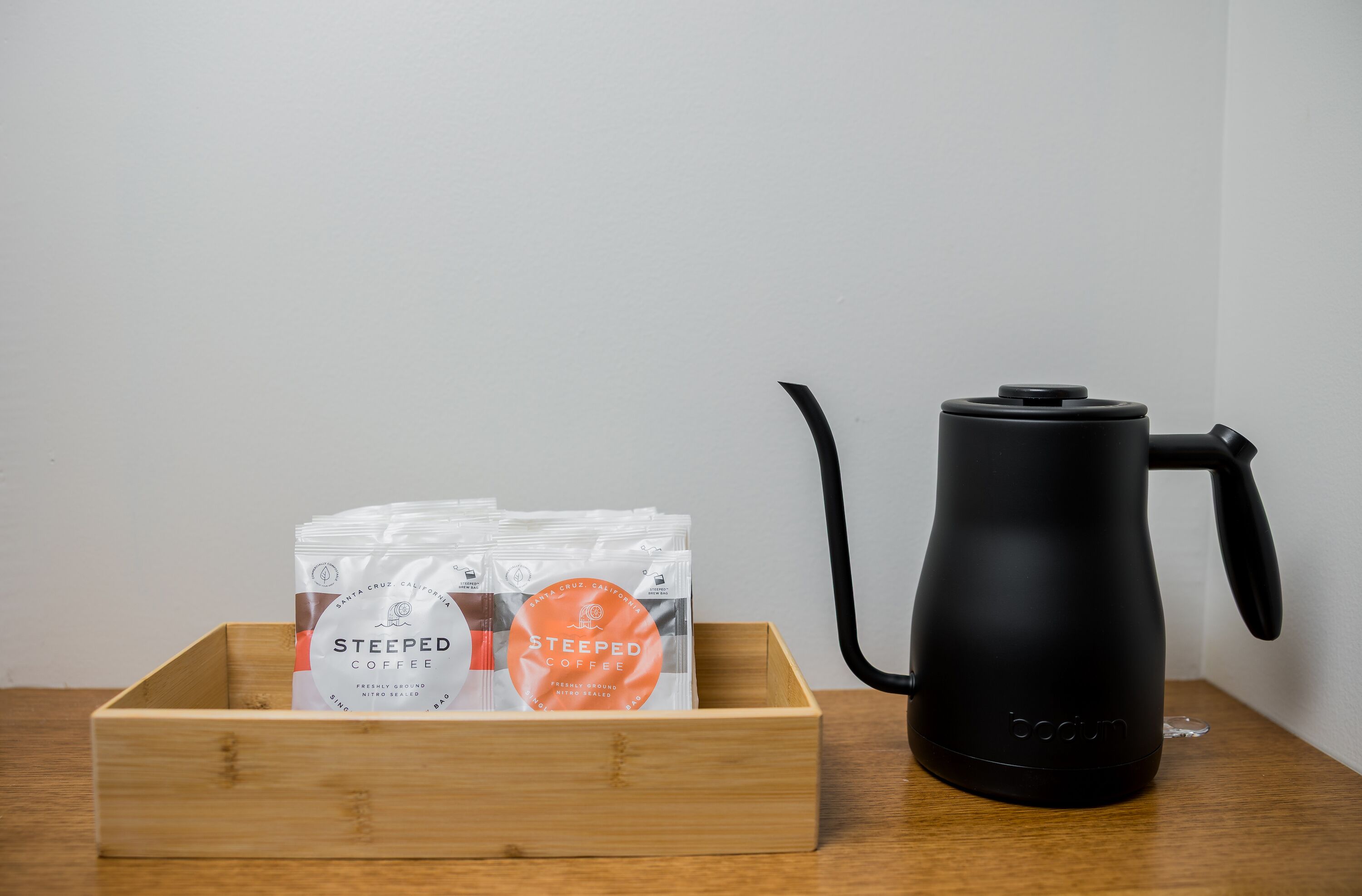 A wooden box containing coffee pods and a black electric kettle on a wooden surface.