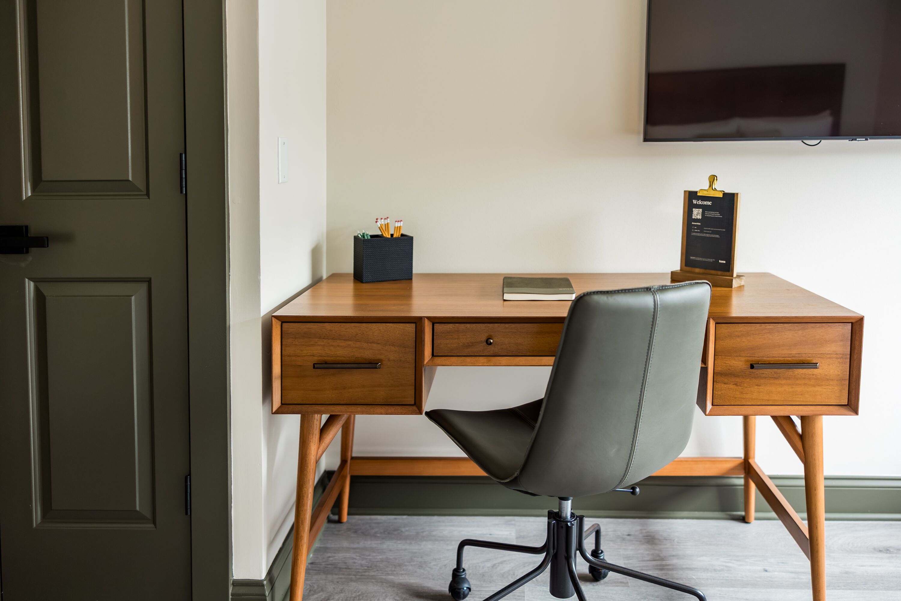 A modern office desk with a leather chair, a flat-screen TV, and a clipboard on the wall.
