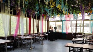 Un restaurant doté d’un plafond haut, décoré de plantes suspendues et de tentures colorées. Des tables et des chaises sont disposées pour accueillir les clients.