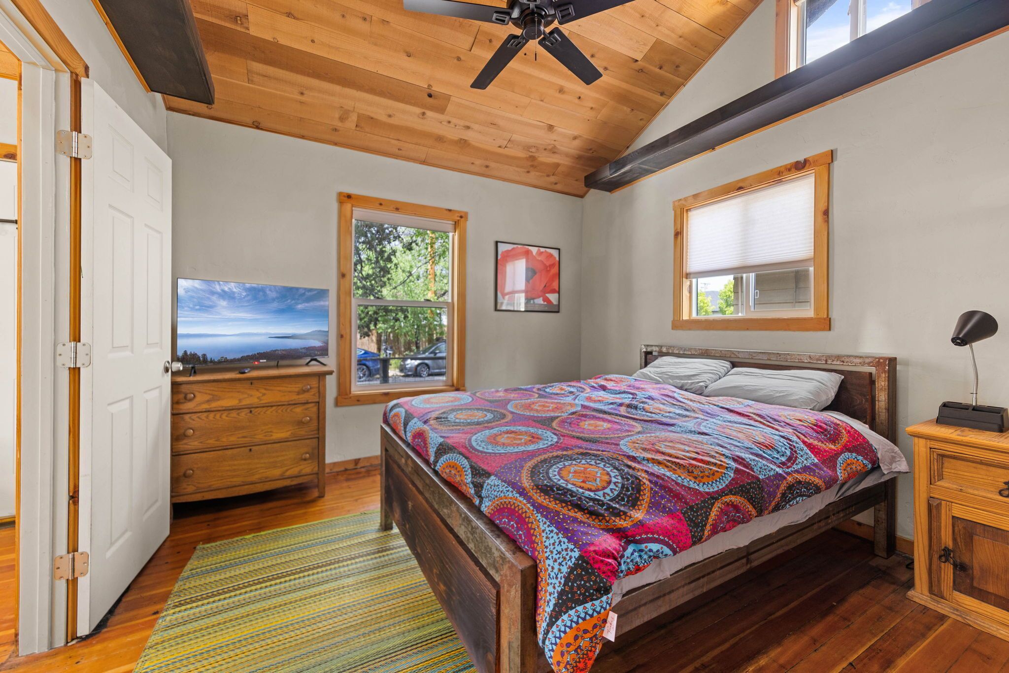 A bedroom with a wooden ceiling, a bed with a colorful blanket, a dresser, a nightstand, a window with a view, and a door.