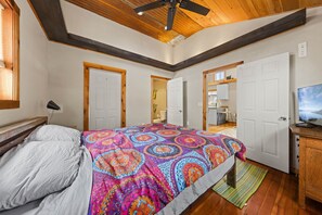 Quarto com cama, ventilador de teto, vigas de madeira e porta que dá para outro cômodo.