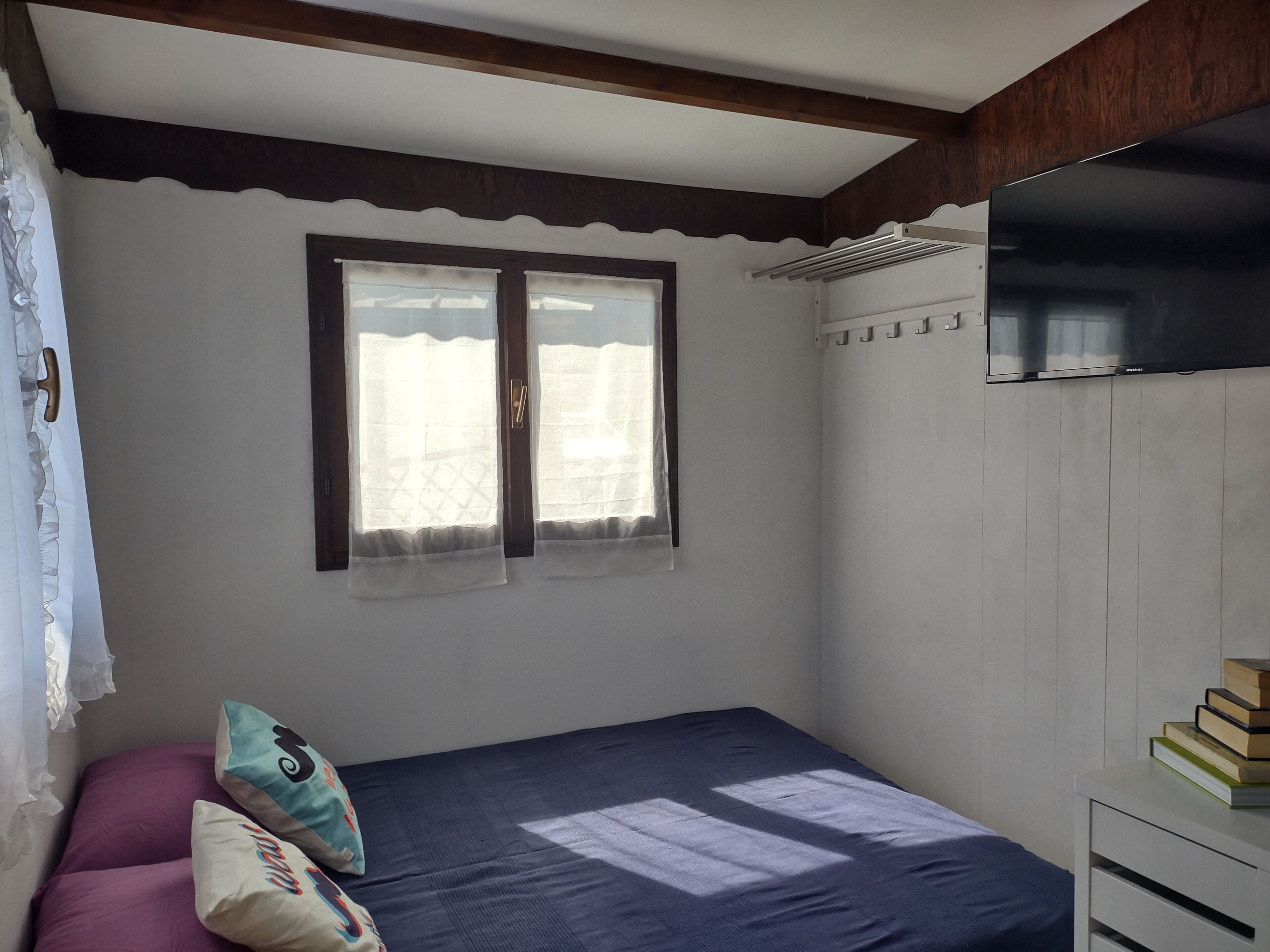 A bedroom with a bed, a window with curtains, a radiator, and a stack of books on a nightstand.