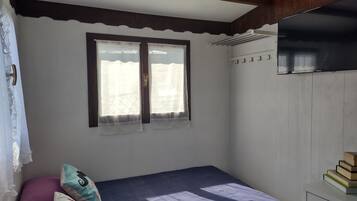 A bedroom with a bed, a window with curtains, a radiator, and a stack of books on a nightstand.