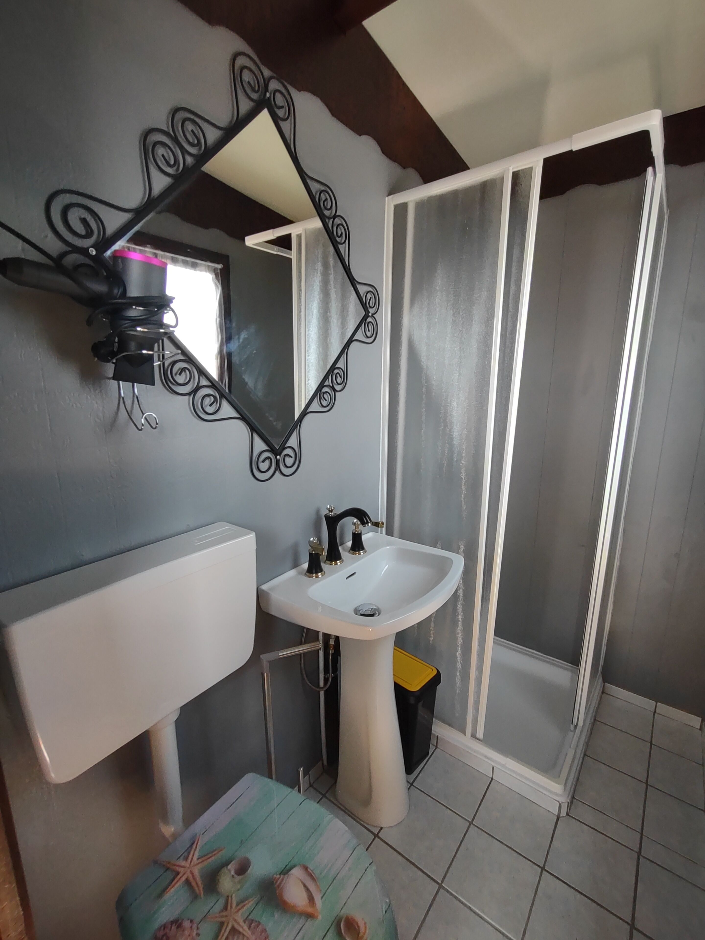 A bathroom with a white sink, a mirror with a decorative frame, a shower enclosure, and a small stool with seashells.