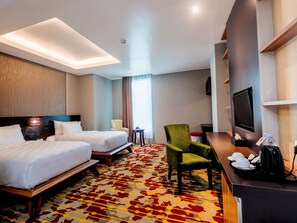 A hotel room with two beds, a green armchair, a desk with a TV, and a patterned carpet. - Grand Sya Hotel (Palu)