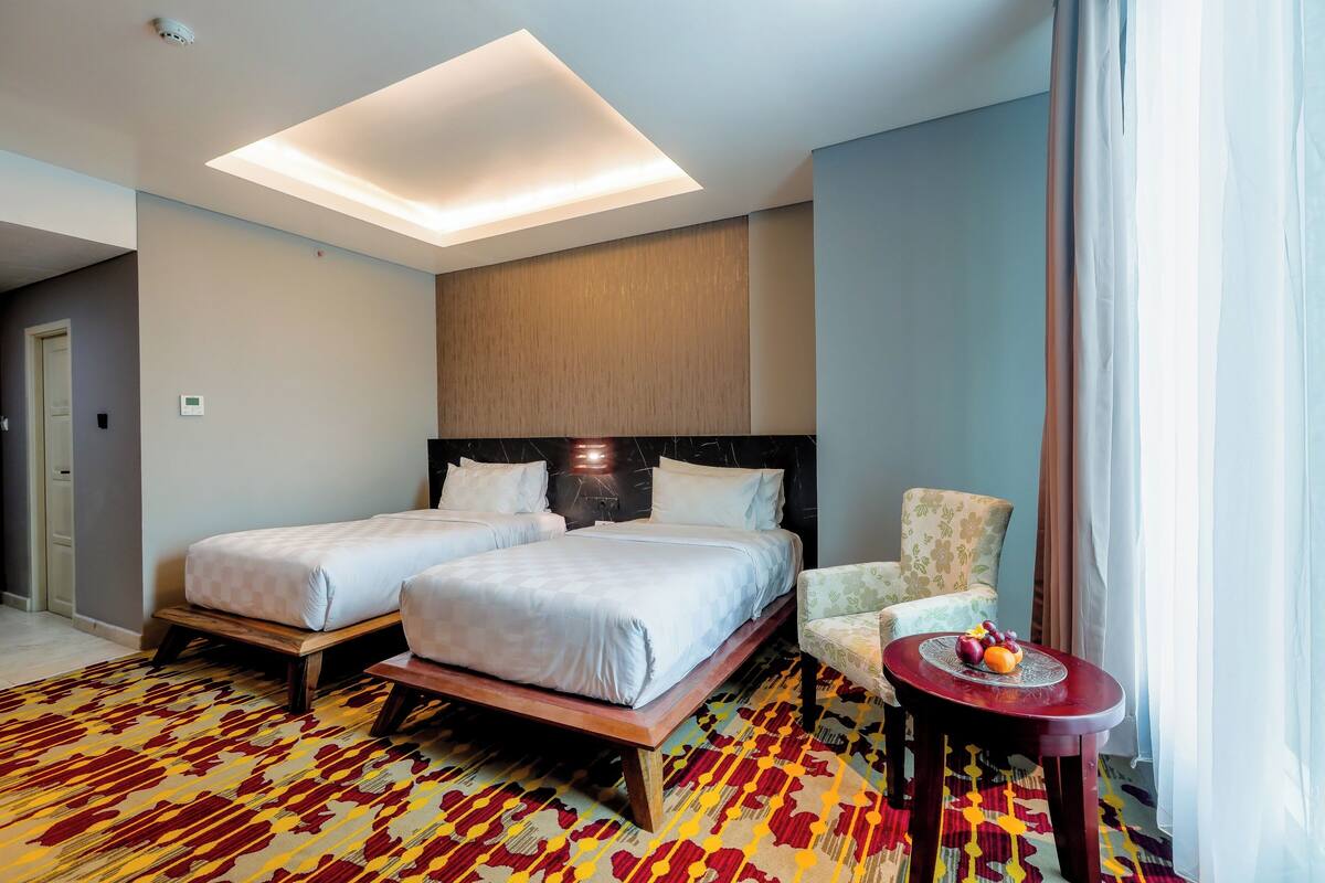 Een hotelkamer met twee bedden, een stoel, een klein tafeltje met fruit en een moderne plafondlamp.