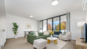 A modern living room with a green sofa, a white armchair, and a coffee table. There is a dining area with a table and chairs in the background.