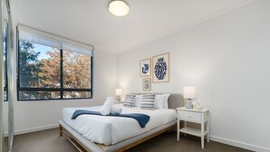 A bedroom with a large window, a bed with white linens and blue accents, a bedside table with a lamp, and two framed artworks on the wall.