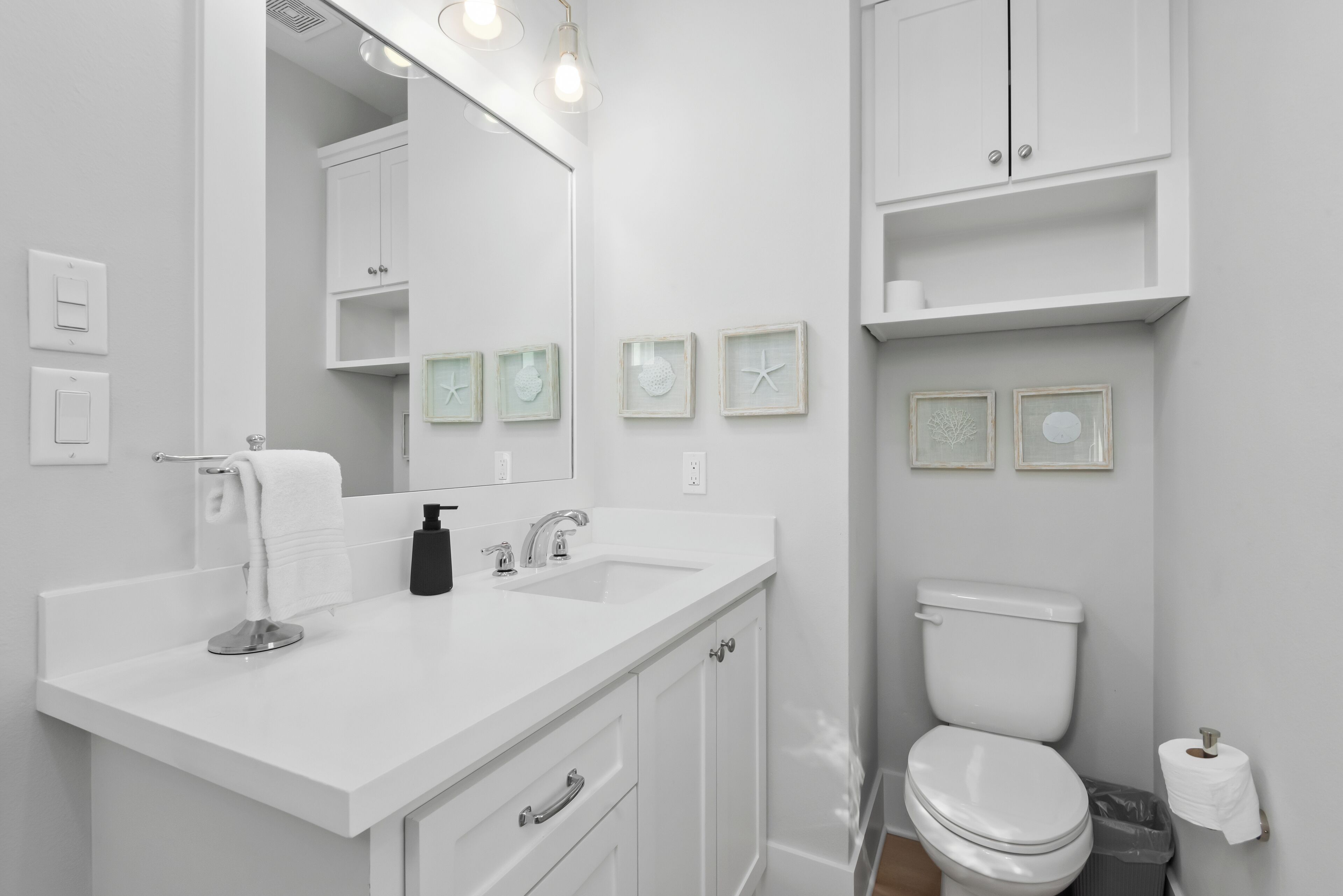 A modern bathroom with a white sink, mirror, and toilet. There are framed pictures on the wall and a trash bin next to the toilet.