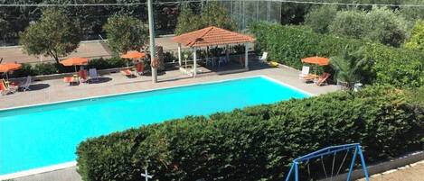 A swimming pool with lounge chairs and umbrellas, surrounded by greenery and a gazebo.