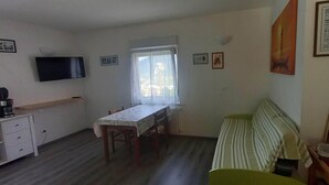 A small, well-lit room with a dining table, a sofa, a TV, and a window with curtains. - Surrounded by nature, cozy apartment with private garden and grill area (Novaledo)