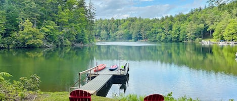A dock extending into a calm lake surrounded by dense forest. Two red Adirondack chairs are positioned on the grassy shore.