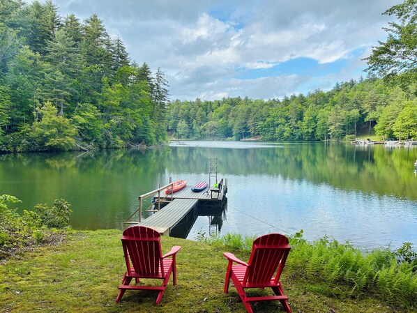 Een steiger die uitsteekt in een kalm meer, omringd door dicht bos. Twee rode Adirondack-stoelen staan op het gras aan de oever.