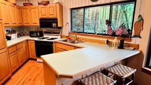A kitchen with wooden cabinets, a stove, a microwave, and a large window with a view of trees. - Treehouse on the Lake! Direct Lakefront Home with Private Dock on Lake Glenville (Cullowhee)