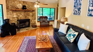 A living room with a stone fireplace, a ceiling fan, a sofa, a coffee table, and a view of trees through large windows. - Treehouse on the Lake! Direct Lakefront Home with Private Dock on Lake Glenville (Cullowhee)