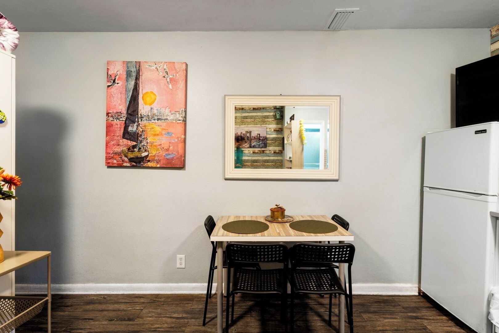 Une cuisine de taille réduite avec un réfrigérateur blanc, une table à manger avec deux chaises et un tableau accroché au-dessus du miroir.