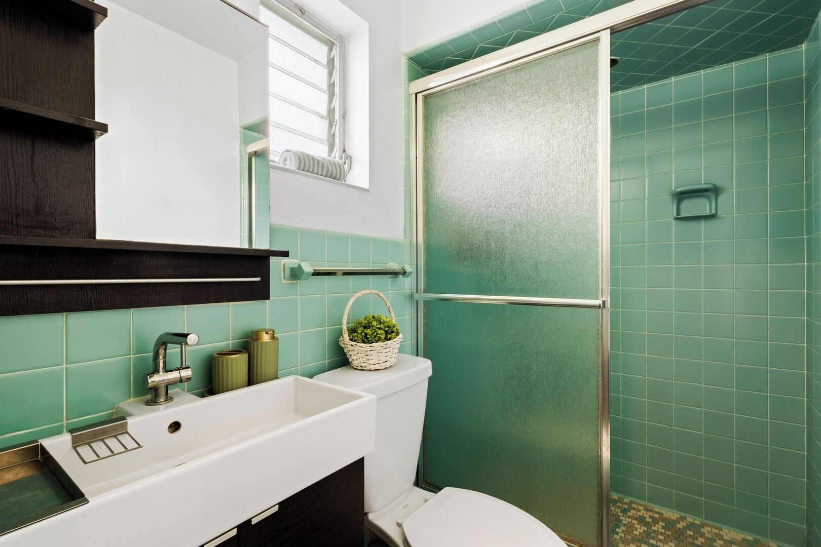 Un bagno con lavandino bianco, uno specchio, una finestra, una doccia con porte in vetro satinato e pareti rivestite di piastrelle verdi.