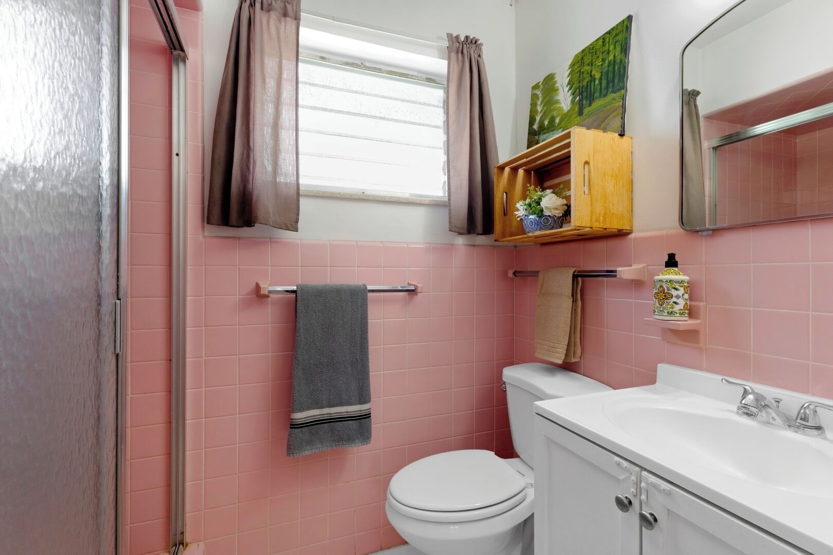 Un baño con baldosas rosas, un inodoro blanco, un lavamanos con espejo y una ventana con cortinas.