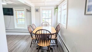 A dining area with a wooden table and chairs, white walls, and large windows.