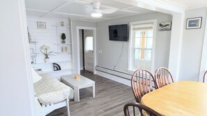 A room with a wooden dining table, chairs, a white sofa, a TV mounted on the wall, and a ceiling fan.