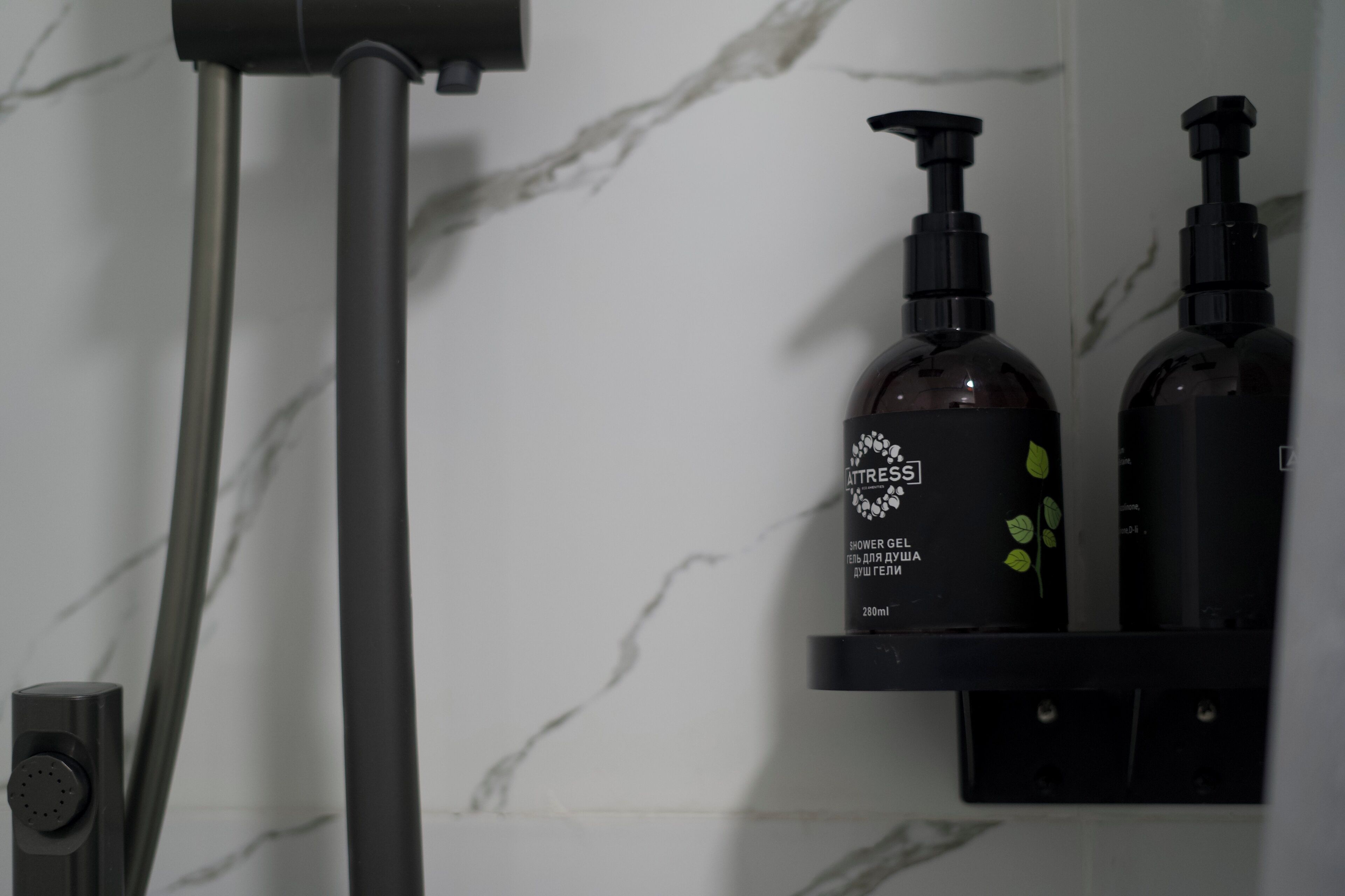 A bathroom with a marble wall, a shower head, and two bottles of shower gel on a black shelf.