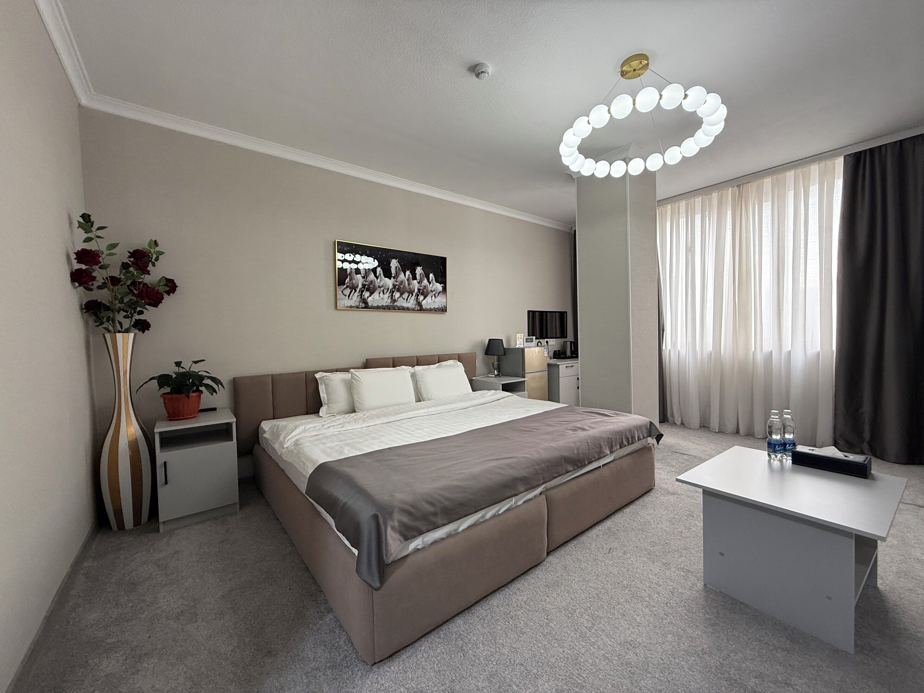 Una habitación de hotel moderna con una cama grande, mesitas de noche, un escritorio, una planta en maceta, un cuadro y un amplio ventanal con cortinas.