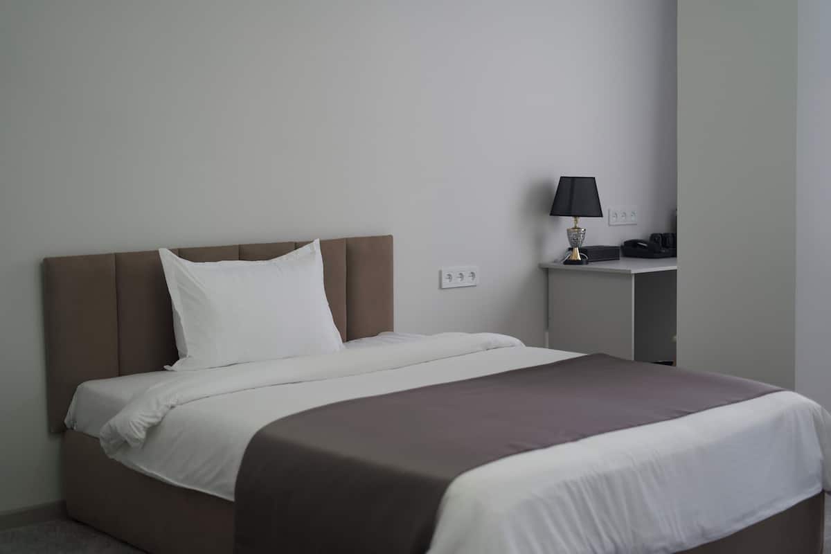 A neatly made bed with white and brown bedding, a small bedside table with a lamp, and a power outlet on the wall.