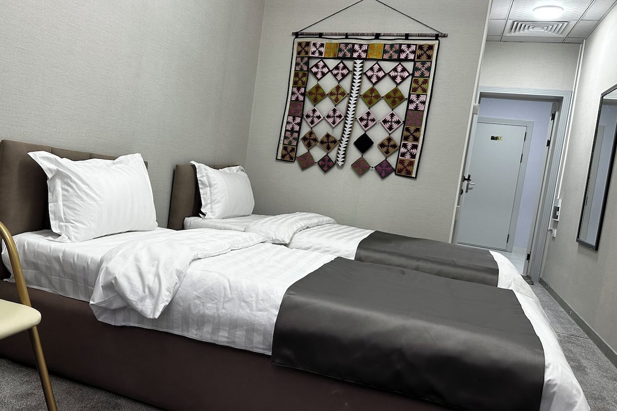 A neatly made bed with white and gray bedding, a brown headboard, and a wall hanging with geometric patterns.