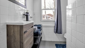 A compact bathroom with white tiles, a wooden vanity, a window with a view of buildings, and a blue bath mat. - Cozy 3BR Near UB & Niagara Falls - Pet Friendly (Buffalo)