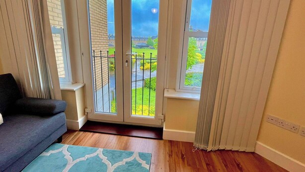 Una habitación con un sofá de color oscuro, una alfombra estampada y vistas a un balcón con césped verde y una pared de ladrillo.