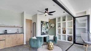 A modern kitchen with wooden cabinets, a countertop, and a ceiling fan. There are two teal armchairs with white cushions, a round gold coffee table, and a white rug. A white shelf with decorative items is visible in the background. - Villa Valena 4 Centrally located, beach, restaurant and promenade within walking distance (Kralendijk)