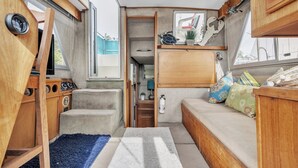 A compact RV interior with a kitchenette, a seating area with cushions, a small window, and a mounted clock. - Luxury Yacht 2 BR Marina Views & Wifi (Anacortes)