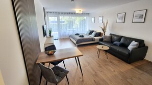 A modern living room with a sofa, a dining table, and a coffee table. There is a large window with a view of greenery outside. - Meerzeit apartment with terrace & parking space (Scharbeutz)