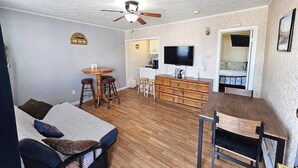 Un espace de vie compact comprenant un lit, une table à manger avec des tabourets, un téléviseur et un ventilateur de plafond.