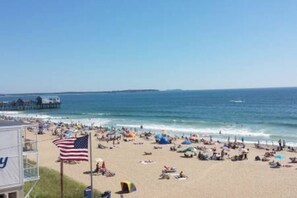 Una playa con un muelle al fondo, una bandera y gente descansando en la arena.