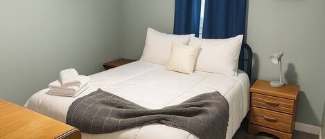 Una cama bien hecha con sábanas blancas, una lámpara de noche y una mesita de madera.