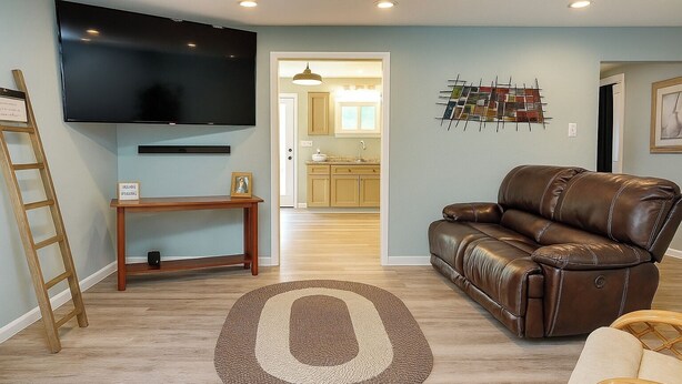 A living room with a brown leather sofa, a wooden coffee table, a TV mounted on the wall, and a kitchen visible through an open doorway. - Cozy 2BR Home: With Lake Access & Work Space (Mercer)