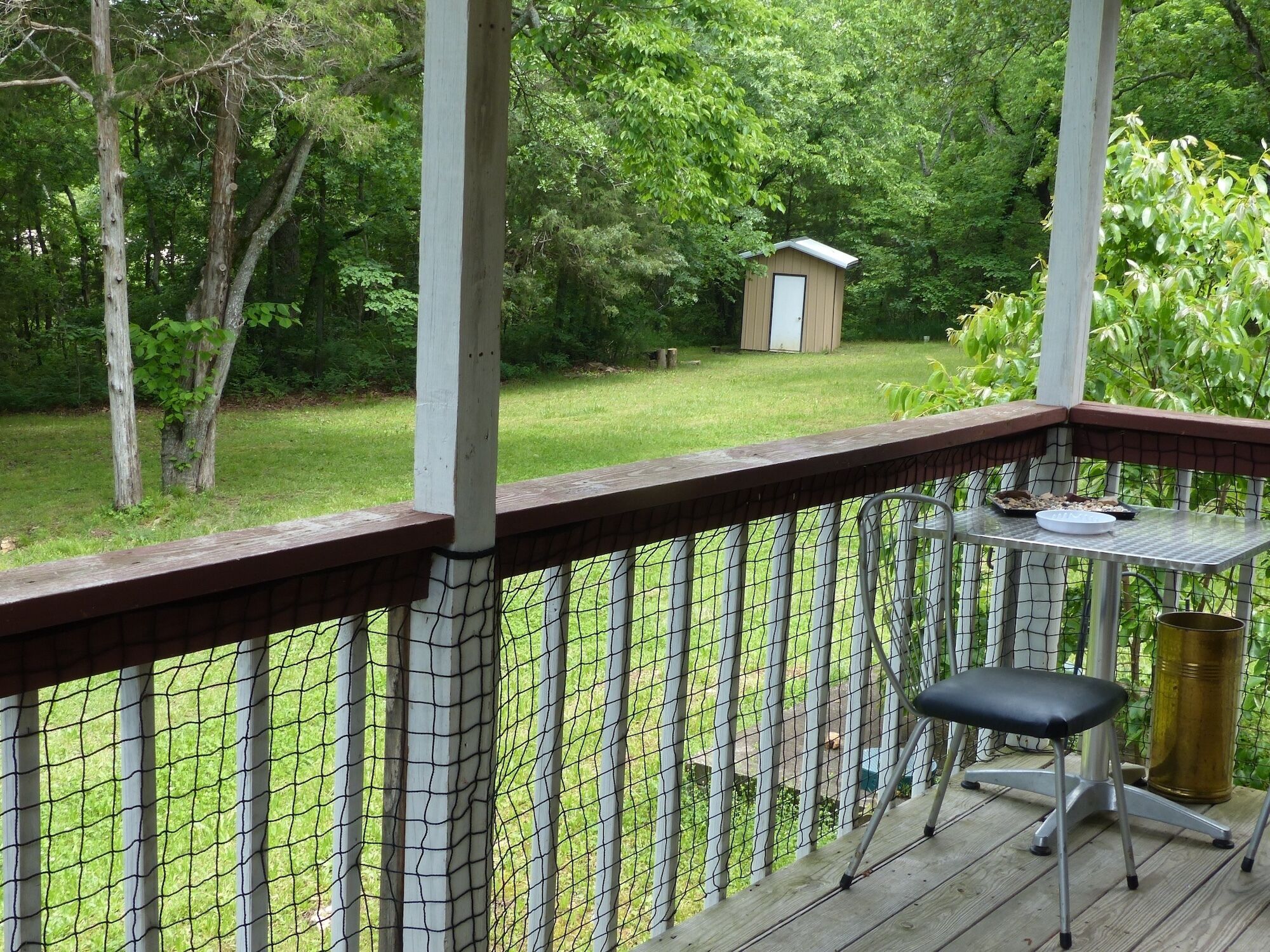 Un espace extérieur abrité, doté d’une terrasse en bois, d’une table en métal avec une chaise, et d’un petit abri de jardin en arrière-plan.