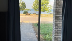 Vista a través de un ventanal hacia un área pavimentada con bancos y un pequeño césped, con vista a un cuerpo de agua y árboles.