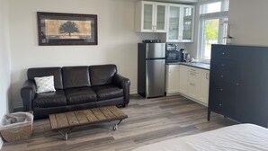 Cocina compacta con refrigerador, microondas y alacenas. Hay una mesa de centro de madera y un sofá de cuero negro con un cojín.