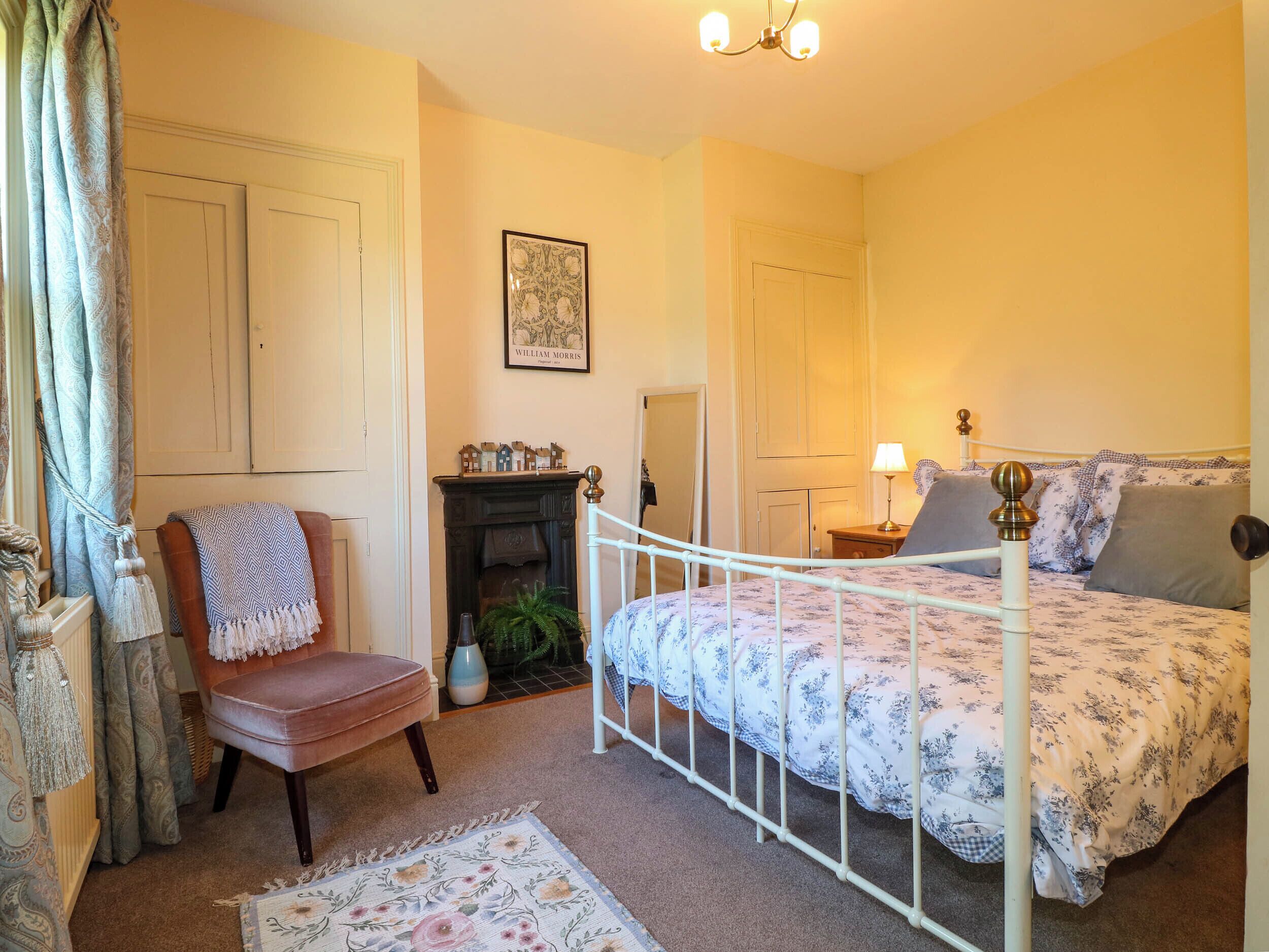 Un dormitorio con cama, una silla, chimenea, lámpara y un cuadro en la pared.