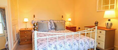 A bedroom with a white metal bed frame, floral bedding, two bedside tables with lamps, a wooden nightstand, and a mirror on the wall.
