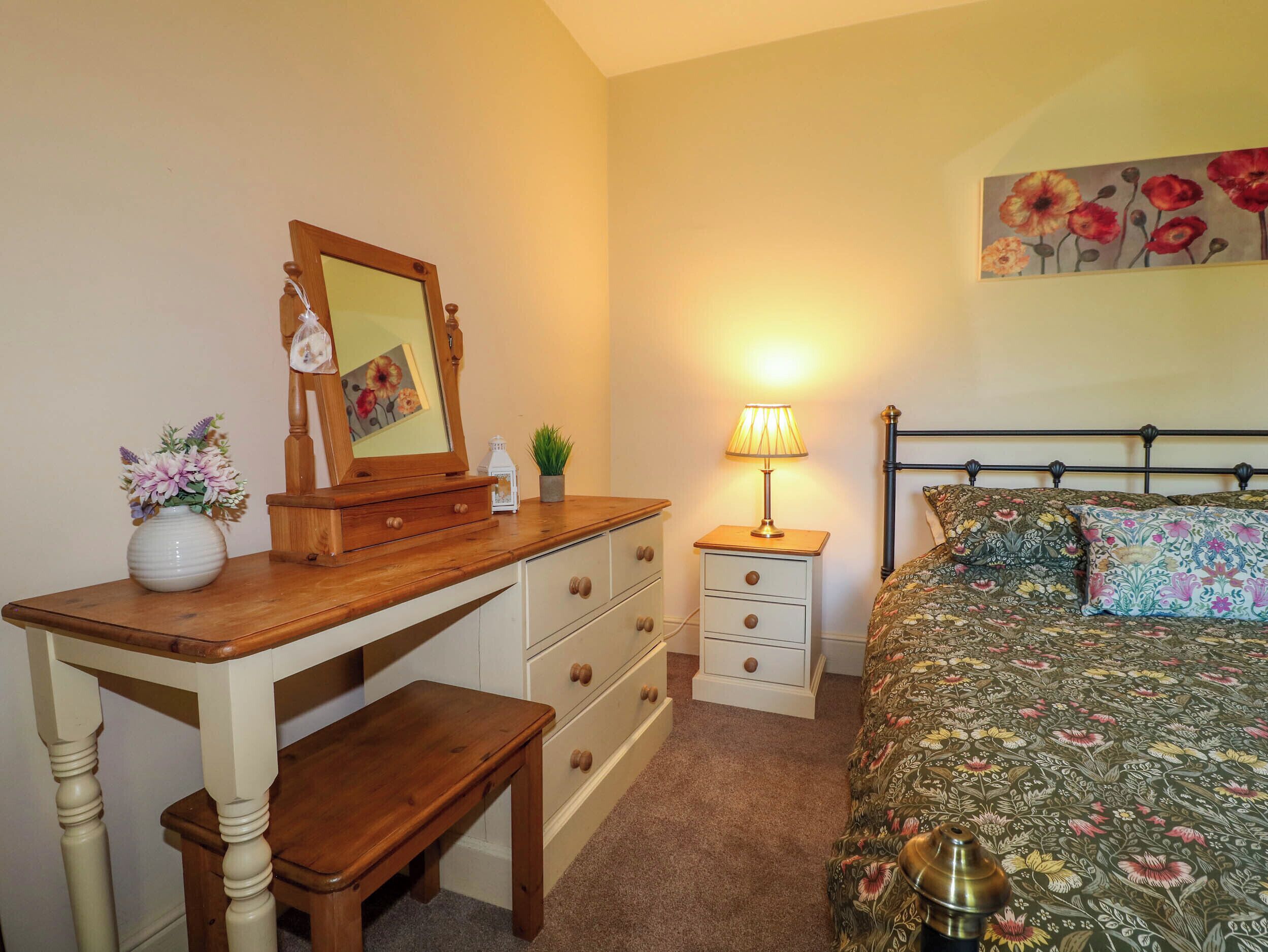 Een slaapkamer met een houten bureau, een bed met bloemenbeddengoed, een nachtkastje met een lamp en een spiegel aan de muur.